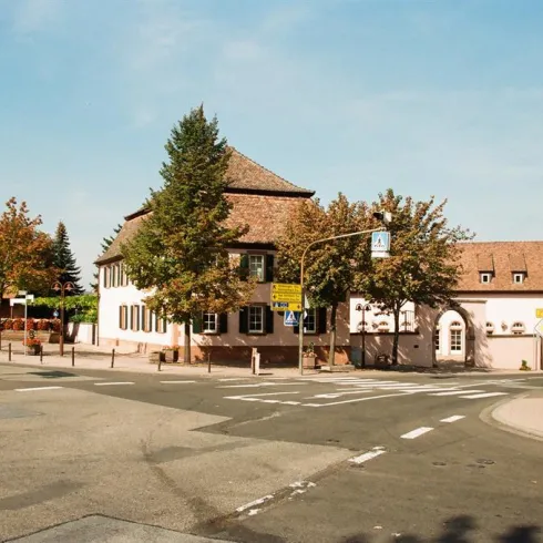 Eine Straßenkreuzung mit mehreren Gebäuden und Bäumen. Der Himmel ist klar und blau.