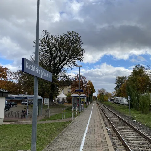 Ein Bahnhof mit Gleisen und einem Wartebereich. Im Hintergrund sind Bäume und Wolken am Himmel zu sehen.
