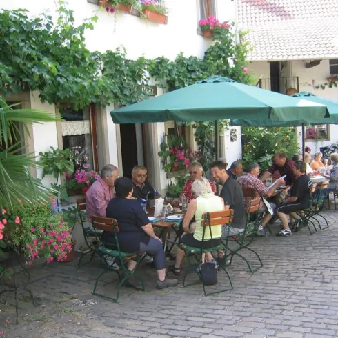 Ein gemütliches Café mit Tischen im Freien. Gäste genießen die Atmosphäre unter einem Sonnenschirm, umgeben von Blumen und Pflanzen.