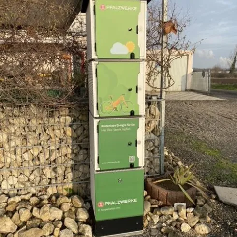 Eine grüne Informationssäule mit Logos und Grafiken, die umweltfreundliche Energie anzeigen. Im Hintergrund sind ein Zaun und einige Pflanzen zu sehen.