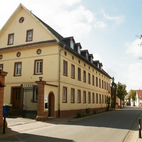 Eine hübsche Straße mit einem historischen Gebäude auf der linken Seite. Die Szene zeigt eine ruhige, städtische Umgebung mit Bäumen und Wohnhäusern.