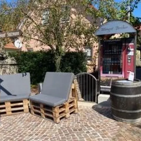 Eine gemütliche Sitzecke mit zwei grauen Stühlen aus Paletten steht auf einem gepflasterten Platz. Neben den Stühlen befindet sich eine Tafel und ein Weinfass.