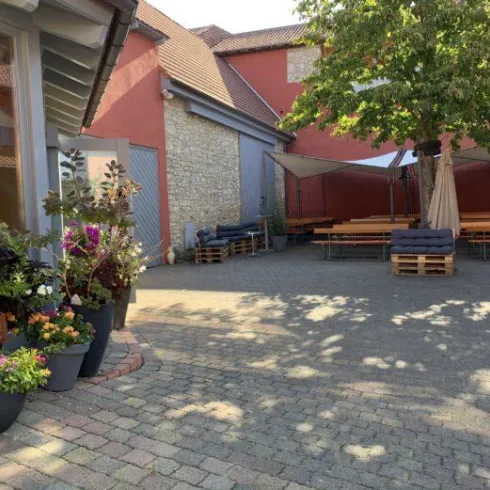 Ein gemütlicher Innenhof mit blühenden Pflanzen und Holzpaletten als Sitze. Im Hintergrund sind Bänke und ein Sonnenschutz zu sehen.