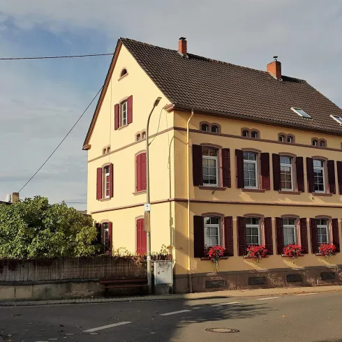 Ein schönes, traditionelles Haus mit gelber Fassade und roten Fensterläden. Die Umgebung ist grün und herbstlich.