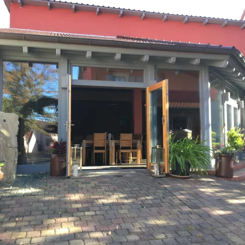 Ein einladender Eingangsbereich mit großen Fenstern und offenen Türen. Im Hintergrund sind Holzstühle und Pflanzen zu sehen.