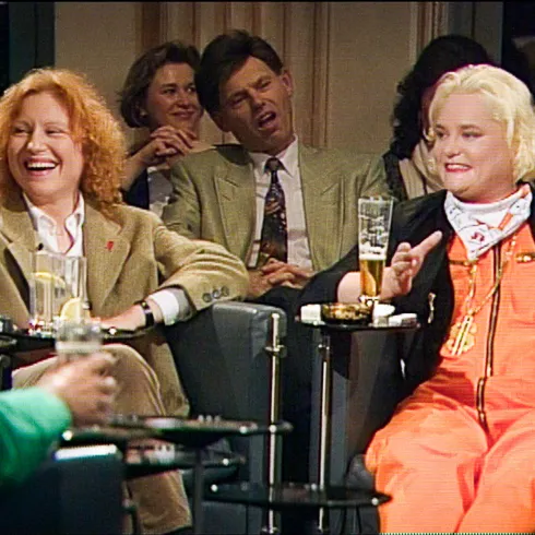 Zwei Frauen lachen und unterhalten sich fröhlich. Im Hintergrund sitzt ein Mann, der scheinbar erschöpft ist.