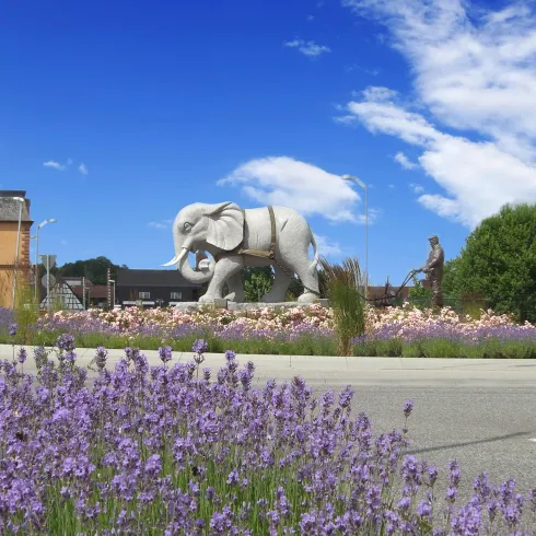 Eine Statue eines Elefanten steht inmitten von bunten Blumen und einem klaren blauen Himmel. Im Hintergrund ist eine kleine Figur zu sehen, die sich neben dem Elefanten befindet.