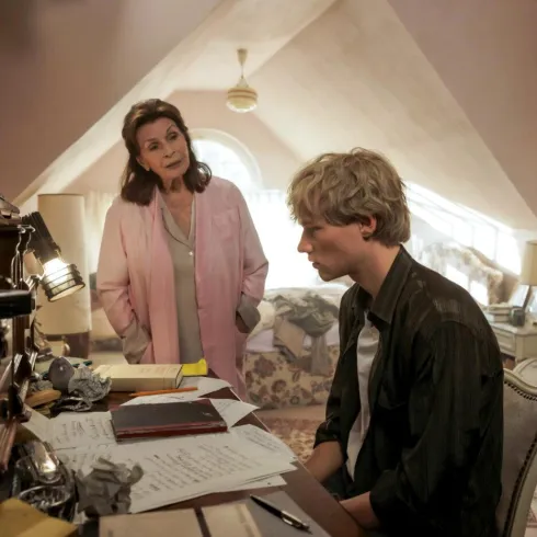 Eine Frau steht neben einem jungen Mann, der an einem Schreibtisch sitzt. Der Raum hat eine gemütliche Atmosphäre mit persönlicher Dekoration.