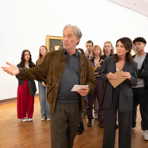 Eine Gruppe von Menschen steht in einem Museum und hört einem Mann und einer Frau zu, die eine Führung leiten. Im Hintergrund sind Gemälde an der Wand zu sehen.
