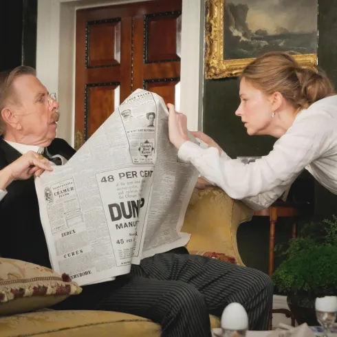 Ein älterer Mann sitzt auf einem Sofa und liest eine Zeitung. Eine junge Frau steht neben ihm und scheint ihn auf etwas aufmerksam zu machen.