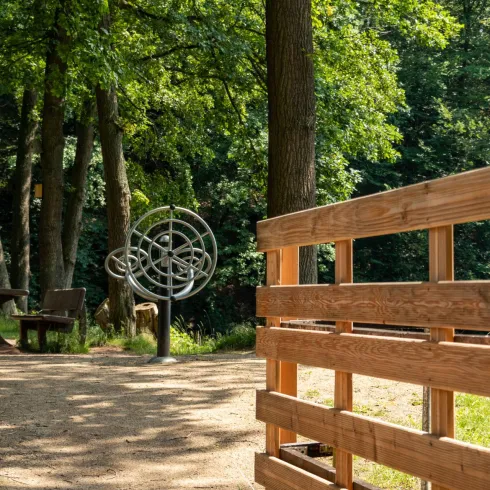 Ein schöner Waldweg mit einem hölzernen Geländer. Im Hintergrund steht eine faszinierende Skulptur zwischen den Bäumen.