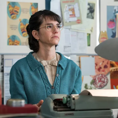 Eine Frau mit Brille sitzt nachdenklich an einem Tisch. Im Hintergrund sind bunte Poster und eine Schreibmaschine zu sehen.