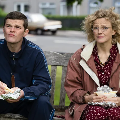 Zwei Personen sitzen auf einer Bank und essen. Der Mann trägt einen Sportanzug, die Frau eine Jacke und blickt nachdenklich.