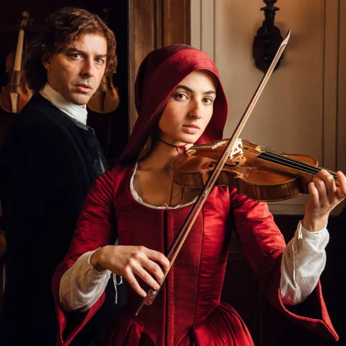 Eine Frau in einem roten Kleid spielt Geige, während ein Mann hinter ihr steht. Die Atmosphäre ist nostalgisch und elegant.