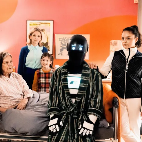 Eine Gruppe von Personen steht in einem Schlafzimmer mit einer älteren Frau im Bett. Ein Roboter in einem Bademantel steht neben ihnen und fügt sich in die Szene ein.