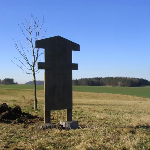 Eine schwarze Informationstafel steht auf einer Wiese vor einem klaren Himmel. Im Hintergrund sind Bäume und ein grünes Feld zu sehen.