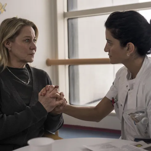 Eine Frau und eine Pflegekraft sitzen sich gegenüber und halten sich an den Händen. Die Atmosphäre ist einfühlsam und unterstützend in einem medizinischen Umfeld.