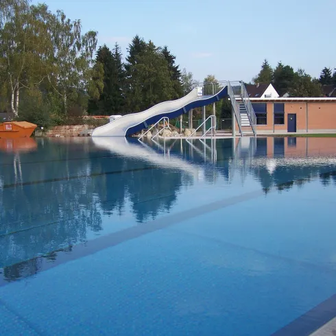 Ein Pool mit einer Wasserrutsche und klar blauem Wasser. Im Hintergrund sind Bäume und ein Gebäude zu sehen.