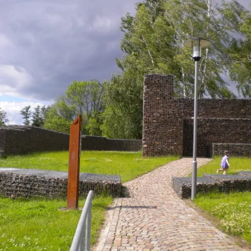 Eine historische Ruine mit einem gepflasterten Weg und einer grünen Wiese. Im Hintergrund sind Bäume und ein grauer Himmel zu sehen.