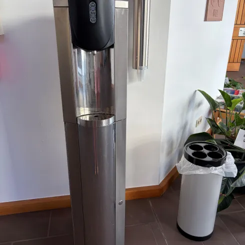 Ein moderner Wasserspender aus Edelstahl steht in einem Raum. Daneben befindet sich ein Abfallbehälter und etwas Grünpflanzen.