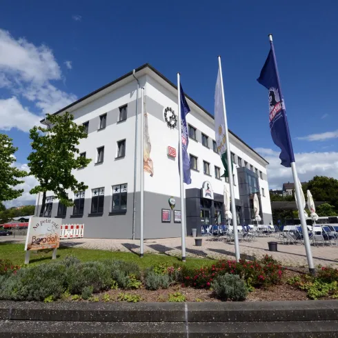 Ein modernes Gebäude mit Fahnen und Grünflächen. Es gibt Bäume und einen klaren Himmel im Hintergrund.