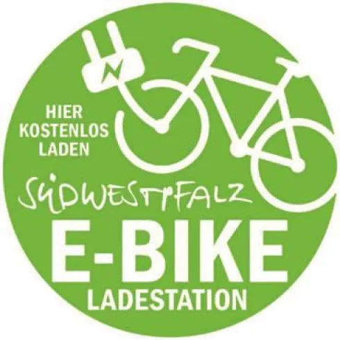 Ein rundes, grünes Schild für eine E-Bike-Ladestation. Es zeigt ein Fahrrad-Symbol und informiert, dass hier kostenlos geladen werden kann.