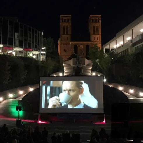 Ein Freiluft-Kino zeigt einen Film auf einer großen Leinwand. Die Kulisse besteht aus stilvollen Gebäuden und einer stimmungsvollen nächtlichen Atmosphäre.