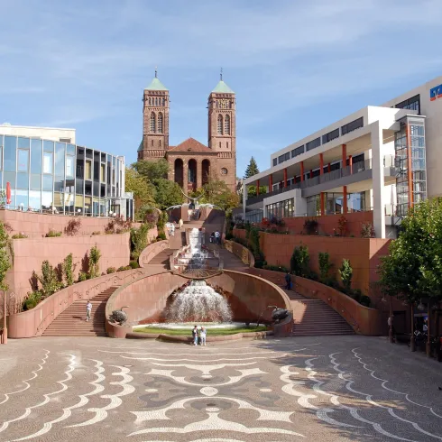 Eine schöne Stadtansicht mit einer Quelle und einem großen Platz. Im Hintergrund sind zwei Türme einer Kirche sichtbar.