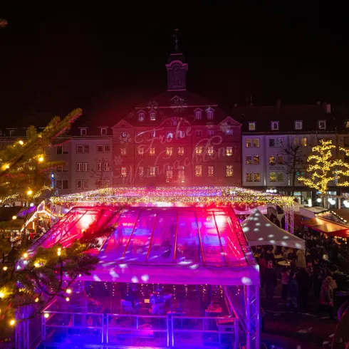 Ein festlicher Weihnachtsmarkt bei Nacht mit bunten Lichtern und geschmückten Ständen. Im Hintergrund ist ein historisches Gebäude mit Lichtprojektionen zu sehen.