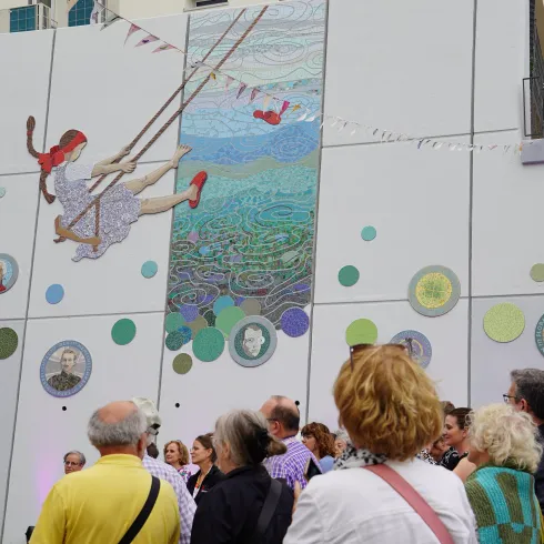 Eine bunte Wandmalerei mit einem schwingenden Mädchen in einem Kleid und roten Schuhen. Vor der Wand sind viele Menschen versammelt, die die Kunst aufmerksam betrachten.