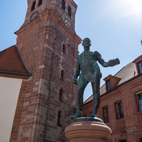 Eine Statue von einem Mann steht vor einem historischen Gebäude mit einem hohen Turm. Der Himmel ist blau und es ist sonnig.
