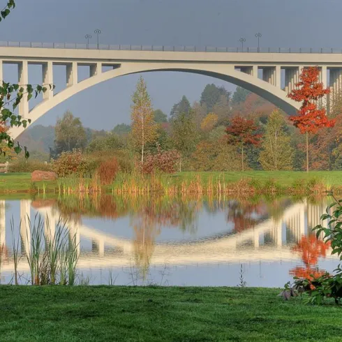 Eine große Brücke mit einem gewölbten Design über einem ruhigen Gewässer. Die Umgebung ist von buntem Laub und grünem Gras umgeben.
