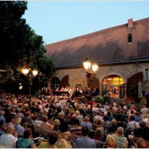 Eine große Menschenmenge sitzt vor einer historischen Halle bei Dämmerung. Im Hintergrund findet ein Konzert statt, das von Laternen beleuchtet wird.