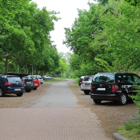 Ein Parkplatz an einem Weg, bei dem beide Seiten mit Autos gefüllt sind. Der Parkplatz ist umgeben von Wald und Bäumen, die Parkfläöchen sind aus Kies.