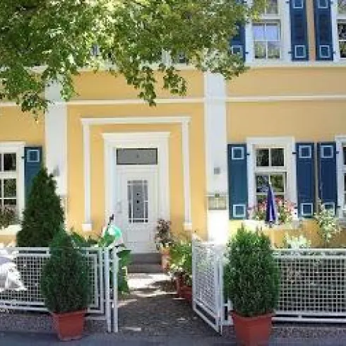 Außenansicht mit Eingangsbereich. Ein schönes gelbes Haus mit blauen Fenstern und einem weißen Zaun. Vor dem Haus stehen grüne Pflanzen in Töpfen.