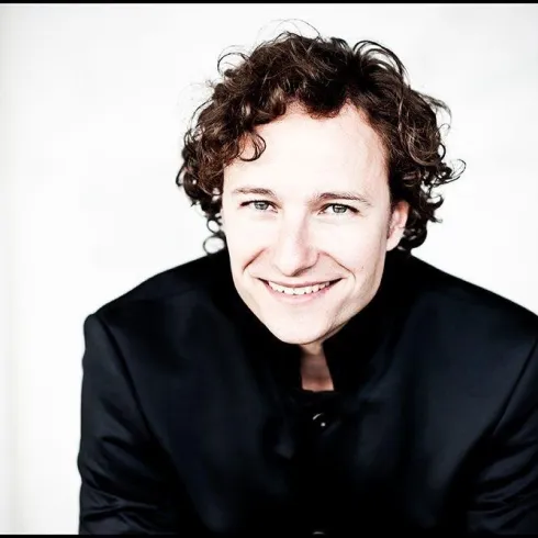 Ein lächelnder Mann mit lockigen Haaren trägt ein schwarzes Outfit. Der Hintergrund ist hell und neutral.