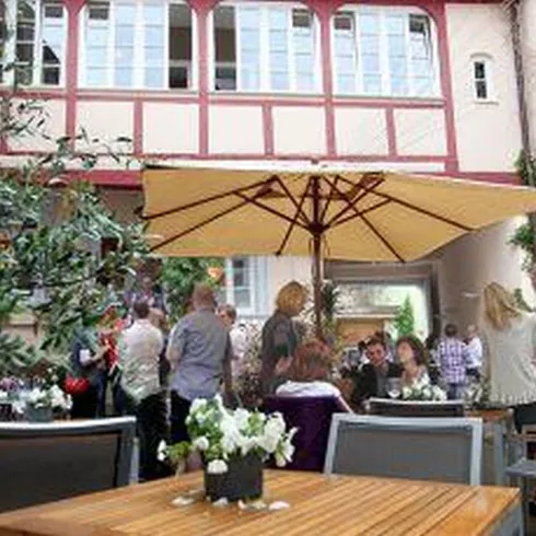 Der Innenhof mit schönen Häusern umgeben. Der Hof ist bestuhlt mit Sitzmöbeln und Sonnenschirmen. Menschen sitzen in diesem Hof und werden von Kellnern bedient.