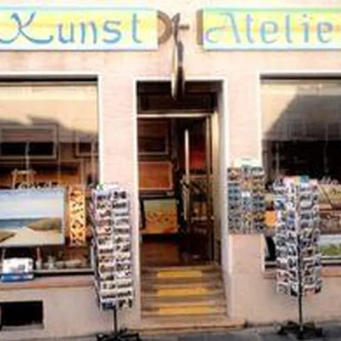 Kunstatelier Seitler. Blick auf die Vorderseite des Hauses mtr zwei  Schaufenstern und zwei Postkartenständern im Außenbereich. In den Schufenstern sind selbstgemalte Bilder von Herrn Seitler ausgestellt.