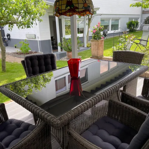 Ein schöner Garten mit einem eleganten Esstisch aus Rattan und bequemen Stühlen. Im Hintergrund sind Pflanzen und eine einladende Sitzecke sichtbar.