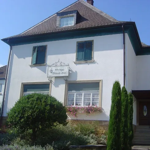 Ein weißes Haus von Vorne. An der Hauswand hängt ein Schild mit dessen Namen. Das Weingut Oßwald - Gaul hat einen Vorgarten, dieser sorgt für eine schöne Atmosphäre.