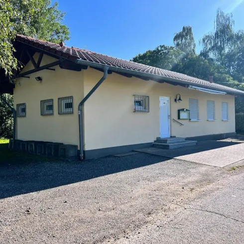 Ein einfaches, gelbes Gebäude mit weißen Fenstern. Es steht an einer Straße, umgeben von Bäumen und Gras.