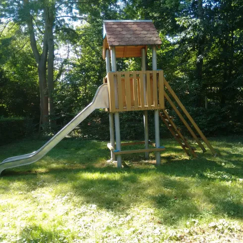Ein Spielplatz mit einem Holzspielgerüst und einer Rutsche. Das Spielgerüst steht in einem grünen Garten, umgeben von Bäumen.