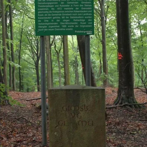 Ein Gedenkstein im Wald mit einer informationalen Tafel darüber. Die Umgebung ist grün und bewaldet.