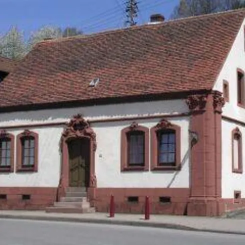 Ein charmantes, historisches Haus mit einem roten Satteldach und dekorativen Fenstern. Es steht an einer Straßenkreuzung in einer ruhigen Umgebung.