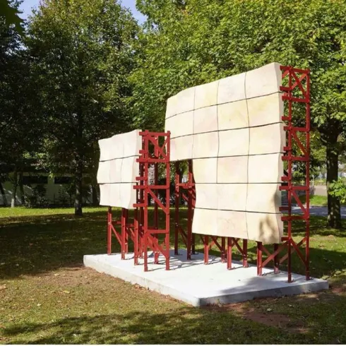 Eine moderne Skulptur aus rotem Metall und hellem Material steht in einem Park. Umgeben von Bäumen bietet sie einen einzigartigen visuellen Kontrast zur Natur.