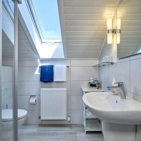 Ein modernes Badezimmer mit Dachfenster und hellem Fliesen. Es enthält ein Waschbecken, eine Dusche und Handtücher in Blau und Weiß.