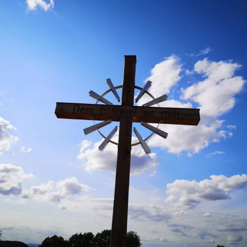 Ein Holzschild in Form eines Kreuzes steht vor einem blauen Himmel mit weißen Wolken. Der Blick ist weit und zeigt eine schöne Landschaft im Hintergrund.