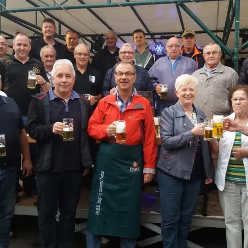 Eine Gruppe von Menschen steht zusammen und hebt Bierkrüge. Sie lächeln und scheinen eine festliche Stimmung zu genießen.