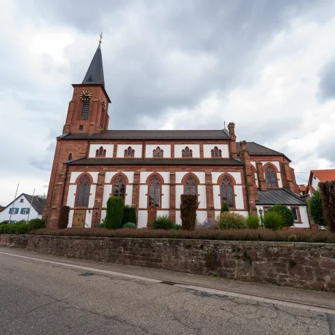 Eine alte Kirche mit einem hohen Turm und bunten Fenstern. Im Hintergrund sind weitere Gebäude und eine bewölkte Himmel zu sehen.