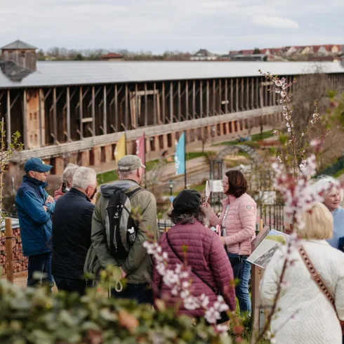 Eine Gruppe von Menschen steht in einem Garten und diskutiert. Im Hintergrund ist ein großes Gebäude zu sehen, und die Pflanzen beginnen zu blühen.